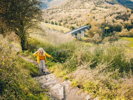 One woman walking a trail in a yellow shirt with a viaduct in the valley below