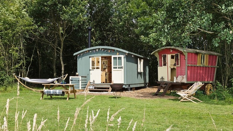 Colourful shepherd's hut and gypsy caravan style accommodation in a tree-edge field with a hammock and picnic table in front at Secret Meadows, Suffolk. Gold winner of the Camping, Glamping and Holiday Park of the Year in the VisitEngland Awards for Excellence 2022
