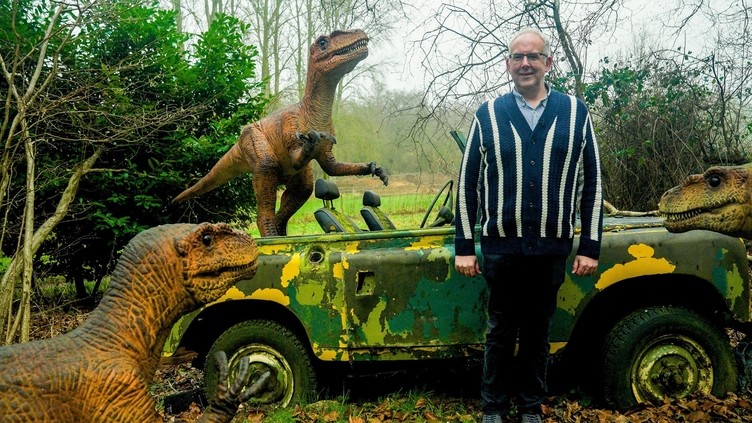 A man poses in front of fake dinosaurs and an camouflaged vehicle