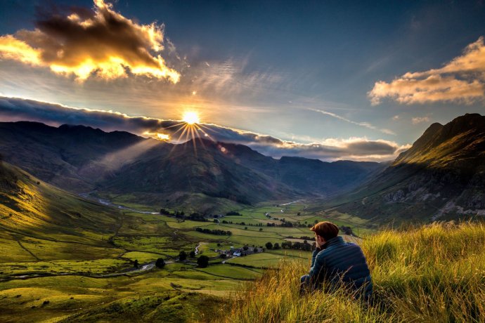 Man sitting in long grass on the side of a fell watching the sunset