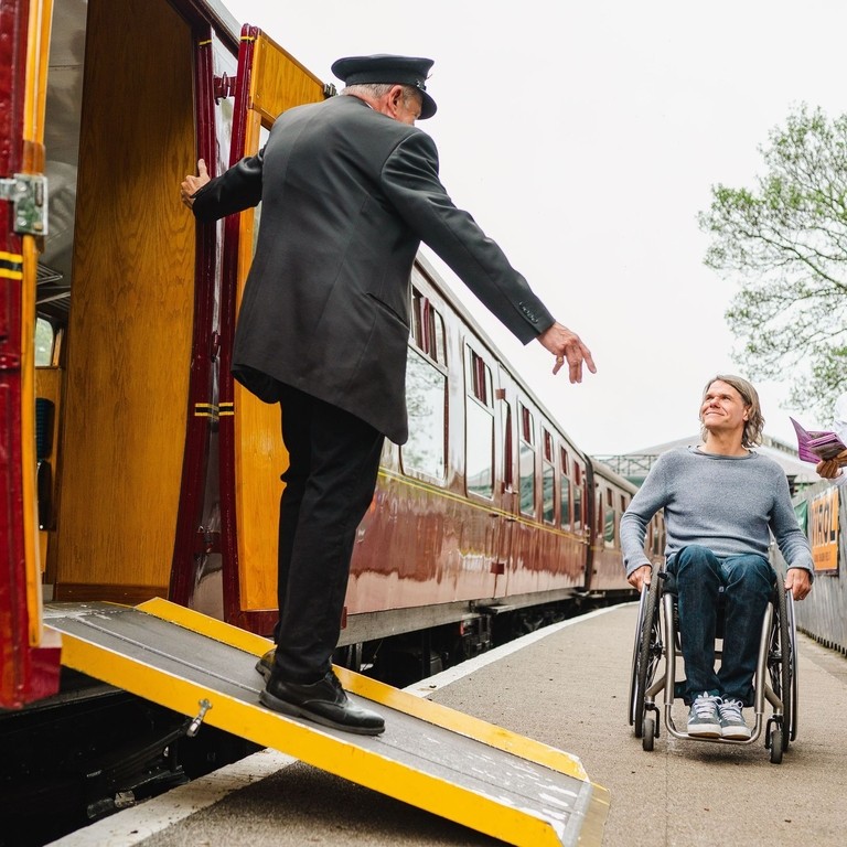 Guard stands on a ramps and leans out of heritage train carriage greeting woman and man using a wheelchair