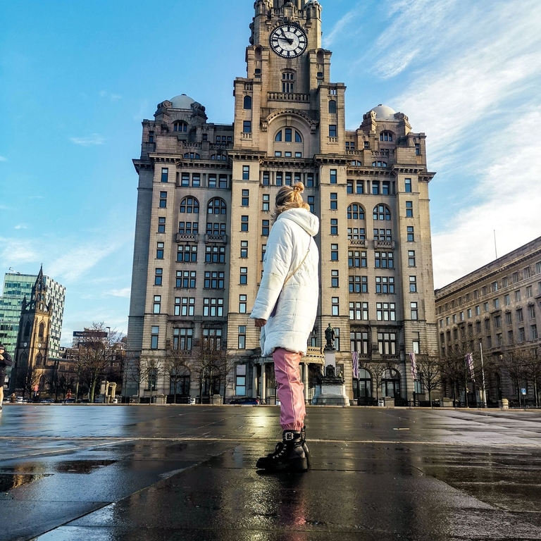 Young woman standing in front of a building in a city centre wearing a white coat.