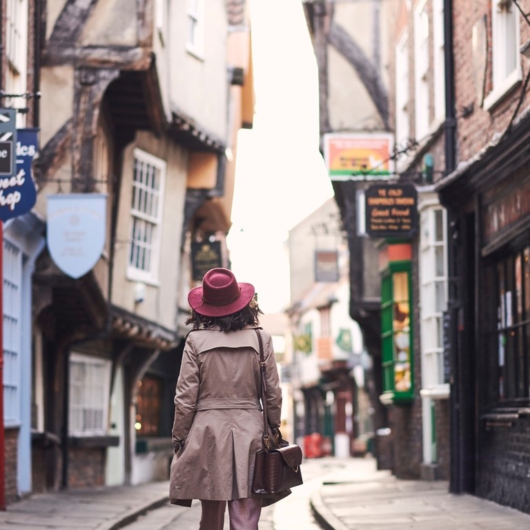 Woman wearing trench coat and pink hat walking through narrow historic street of York, North Yorkshire, England.