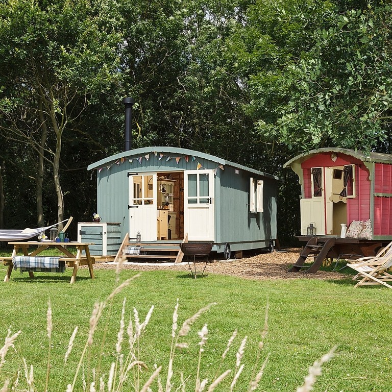 Colourful shepherd's hut and gypsy caravan style accommodation in a tree-edge field with a hammock and picnic table in front at Secret Meadows, Suffolk. Gold winner of the Camping, Glamping and Holiday Park of the Year in the VisitEngland Awards for Excellence 2022