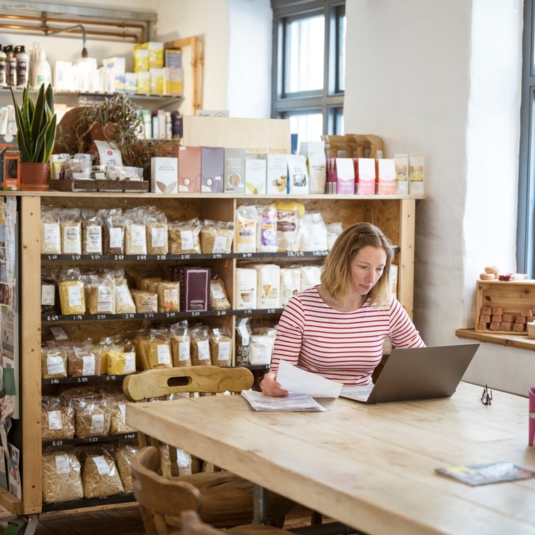 Caucasian businesswoman in early 40s using laptop to sort out paperwork while sitting at communal table in her health food shop and cafe