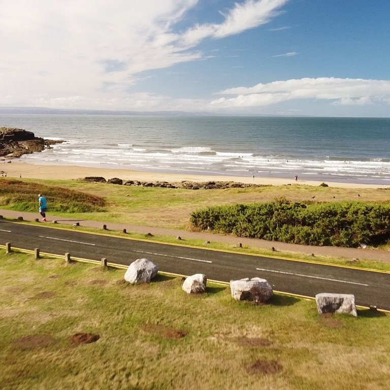 A Campervan drives along a coastal road in front of a Porthcawl Beach.