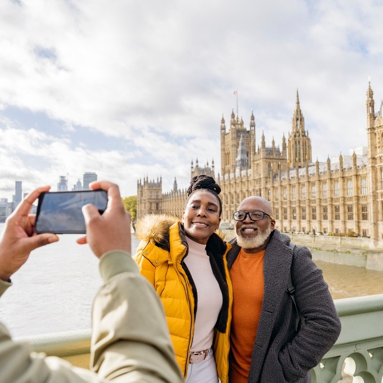 A man photographing mature couple with River Thames and Houses of Parliament in background.