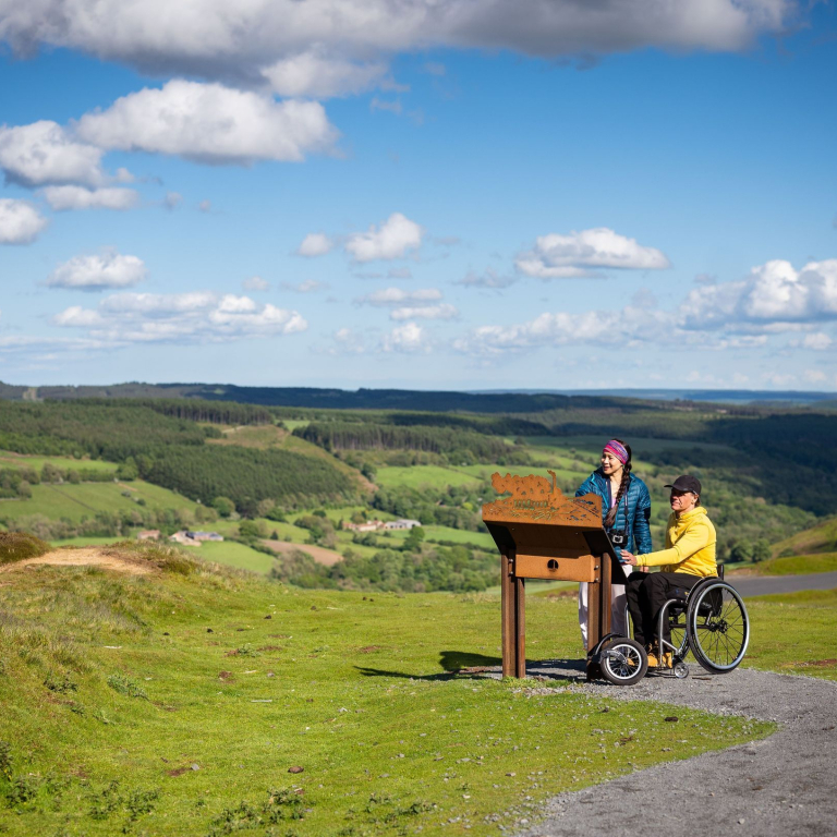 Man and woman at the top of a hill and the man is using a wheelchair and binoculars