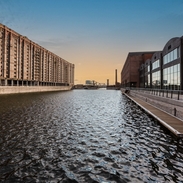 A view of a dock looking across the water. To the left stands a traditional brick built dockside building, to the right is a modern building made from glass and wood. 