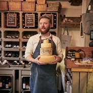 Man wearing blue apron holding a stack of cheeses in a cheese and wine shop