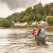 Two people canoeing down a river through a countryside town.