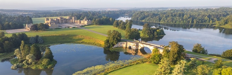 An aerial view of the grounds surrounding Blenheim Palace, an eighteenth-century country house, surrounded by gardens, trees and a lake crossed by a bridge to reach the front of the house. Blenheim Palace - Silver award winner for the Large Visitor Attraction of the Year at the VisitEngland Awards for Excellence 2023.