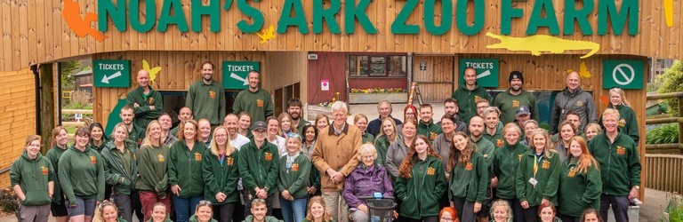 A sign which reads "Welcome to Noah's Ark Zoo Farm" with various staff members standing in front wearing matching branded tops. Noahs Ark Zoo Farm - Gold award winner for the Accessible and Inclusive Tourism Award at the VisitEngland Awards for Excellence 2023.