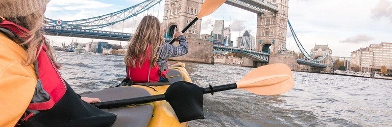 Two women paddling in a kayak on a river towards a bridge in a city