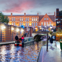 Riverboat cruising down a canal at dusk, with people walking down a parallel path.