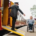 Guard stands on a ramps and leans out of heritage train carriage greeting woman and man using a wheelchair