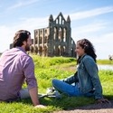 Man and woman sitting on grass with crutches beside them, Whitby Abbey in the background