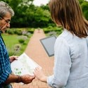 Rear view of male gardener sharing garden plans with female garden designer holding an iPad stood on a path in a garden