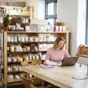 Caucasian businesswoman in early 40s using laptop to sort out paperwork while sitting at communal table in her health food shop and cafe