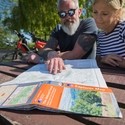 Two people sat at a picnic table, looking at an Ordinance Survey Explorer map of the National Forest. Two Ebikes are parked behind them. National Forest EBike Holidays - Silver award winner for the Ethical, Responsible and Sustainable Tourism Award at the VisitEngland Awards for Excellence 2023.
