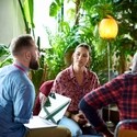 Two men and two women are sitting in a circle of chairs talking in front of a backgdrop full of green leafy plants and one orange glowing floor lamp. One man is an arm amputee and has a laptop on his legs.